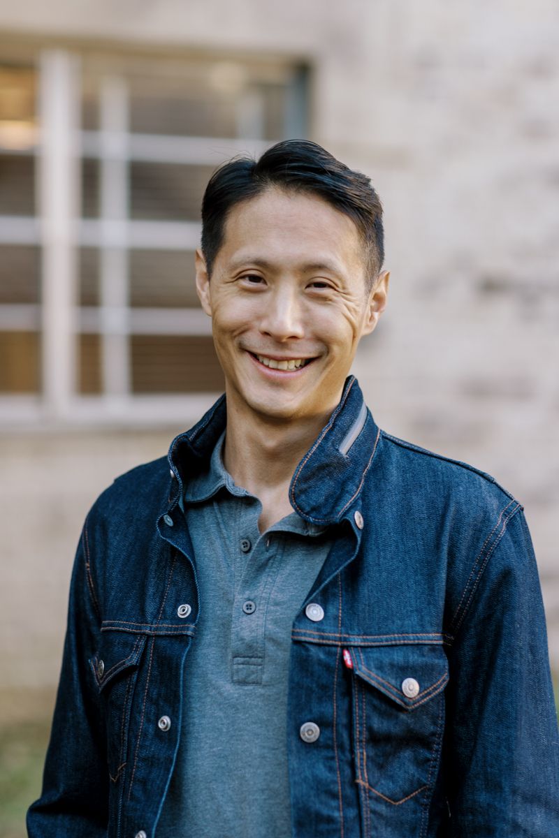 Smiling Asian man in denim jacket standing outdoors in front of a building on a sunny day.