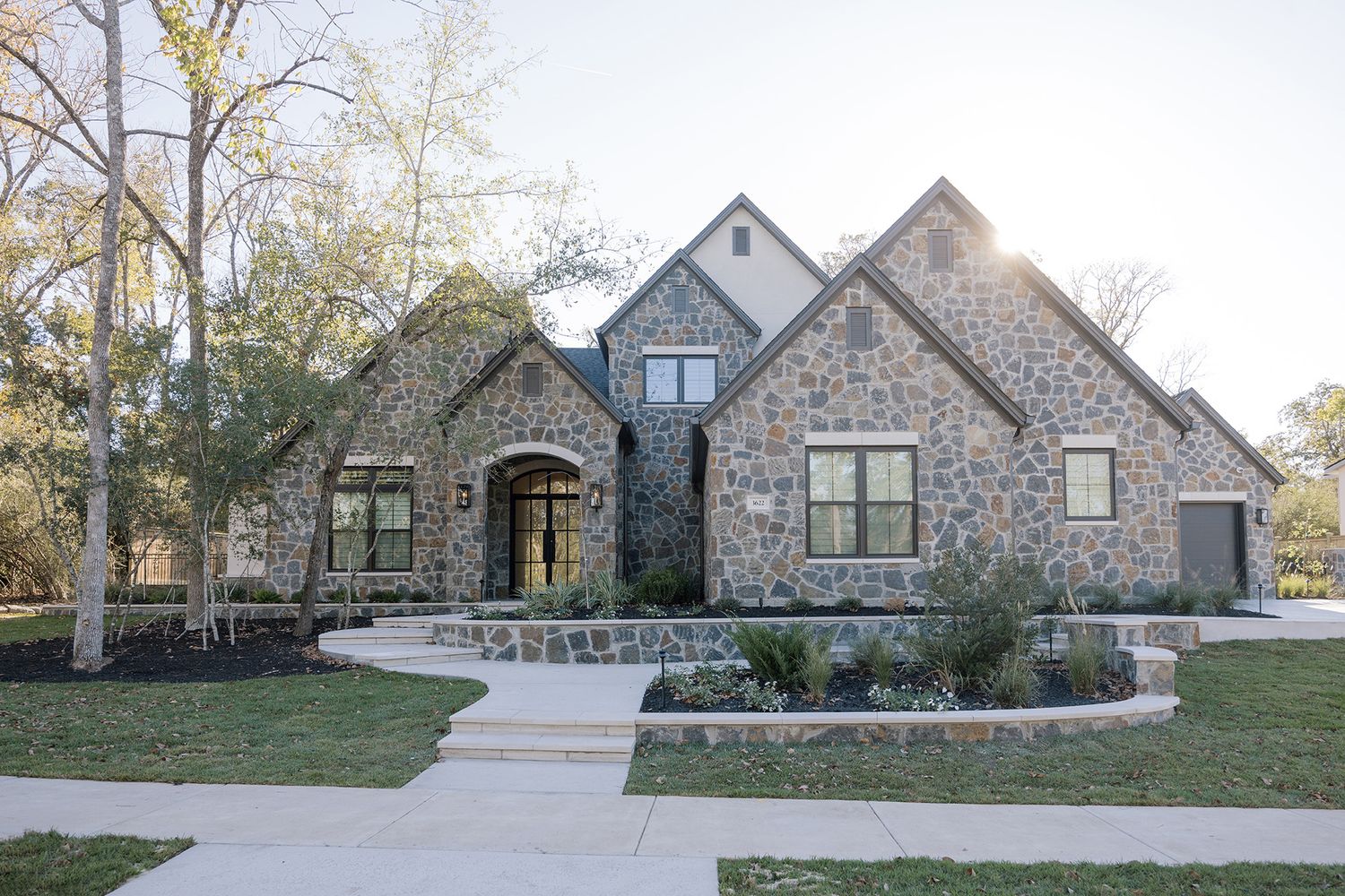 Stone and brick luxury home exterior with arched entry, manicured landscaping, and large windows at sunset.