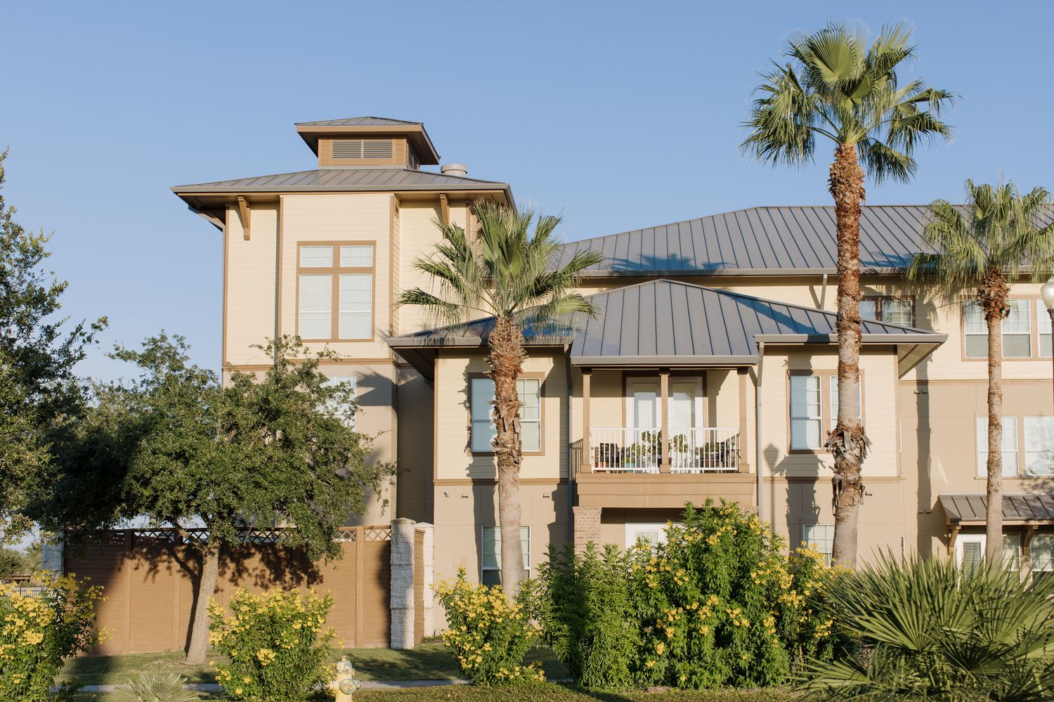 Luxury three-story beige stucco home with palm trees, metal roof, and balconies surrounded by lush landscaping.