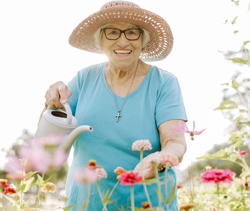 Smiling senior woman in straw hat and glasses watering colorful garden flowers on a bright sunny day.