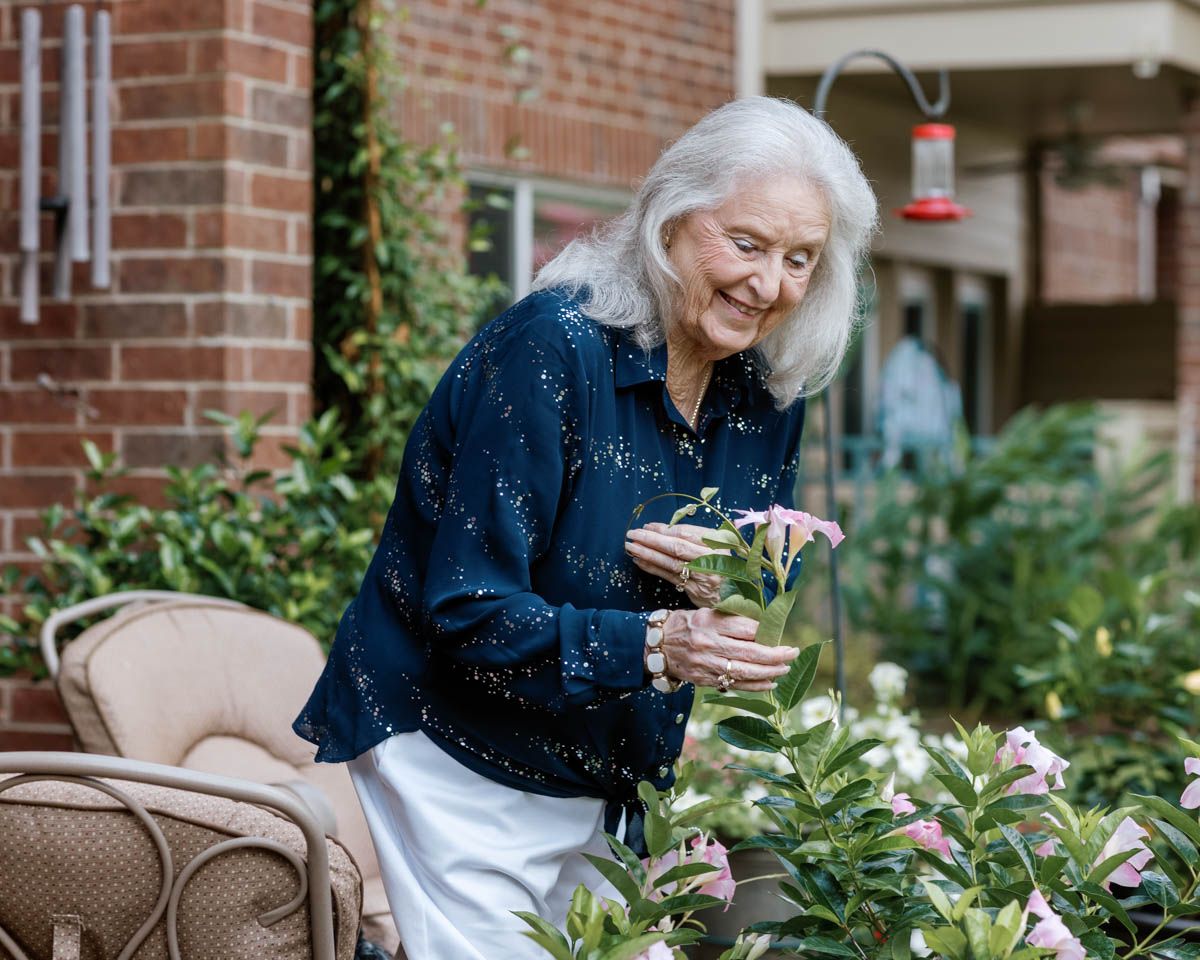 Elderly woman with white hair smiling while tending to pink flowers in a brick courtyard garden.