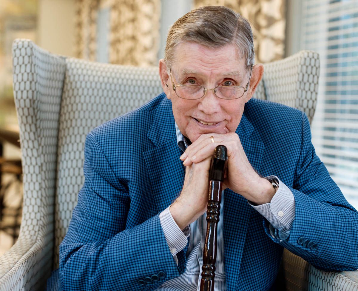 Elderly man in blue blazer sitting in chair, leaning on wooden cane with thoughtful expression.