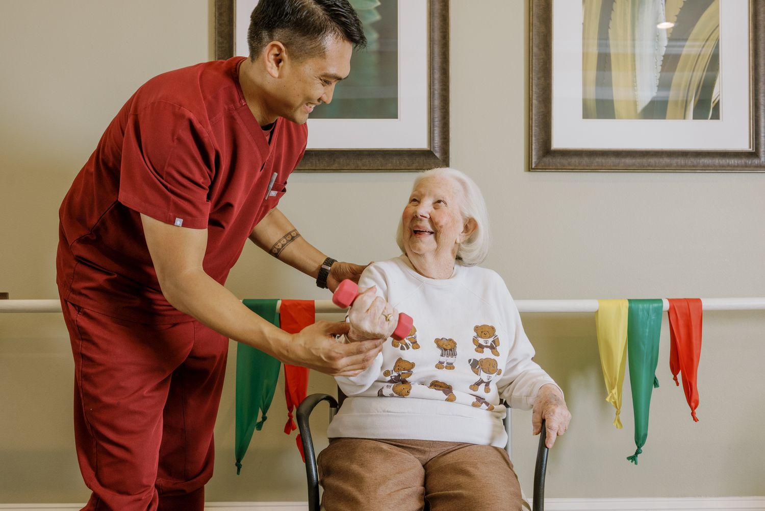 A caregiver in red scrubs smiles while handing a gift to an elderly woman seated in a chair at a care facility.