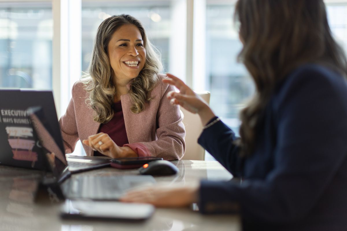 Two professional women in business attire having a friendly meeting at an office desk.