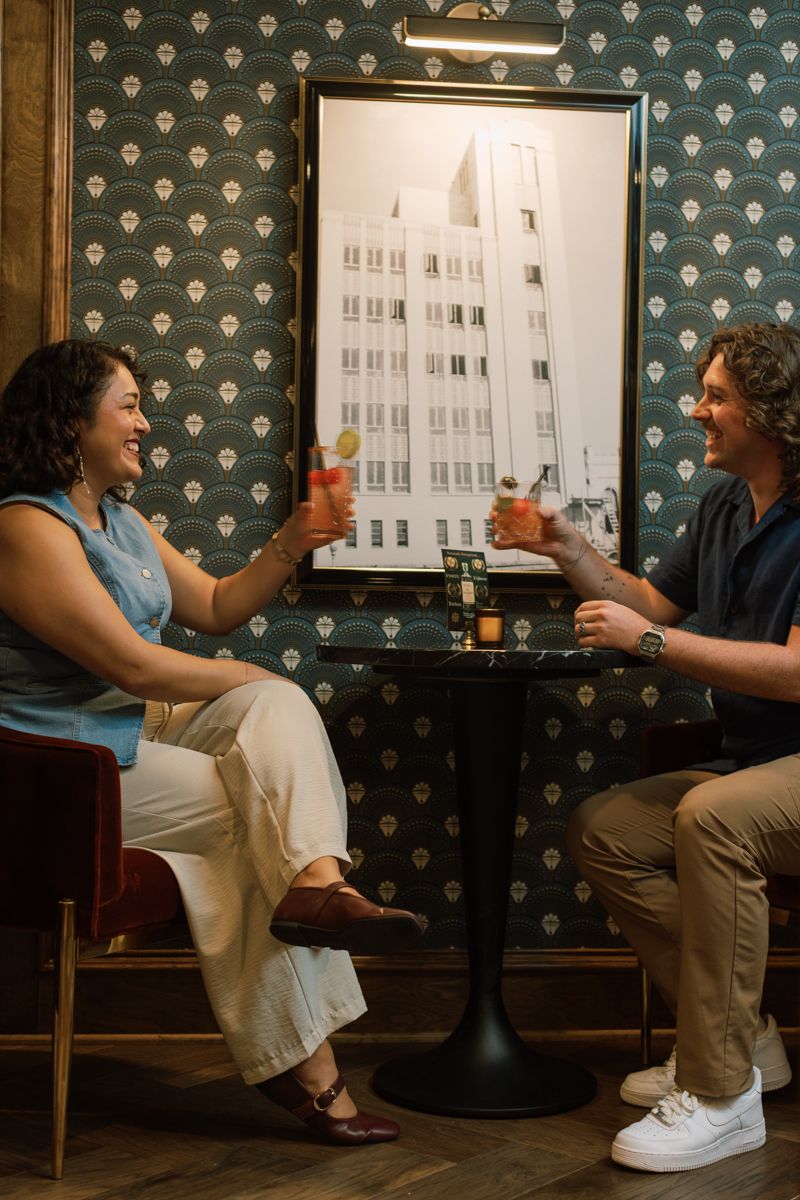 A man and woman toast cocktails at a small table in a stylish bar with art deco wallpaper.