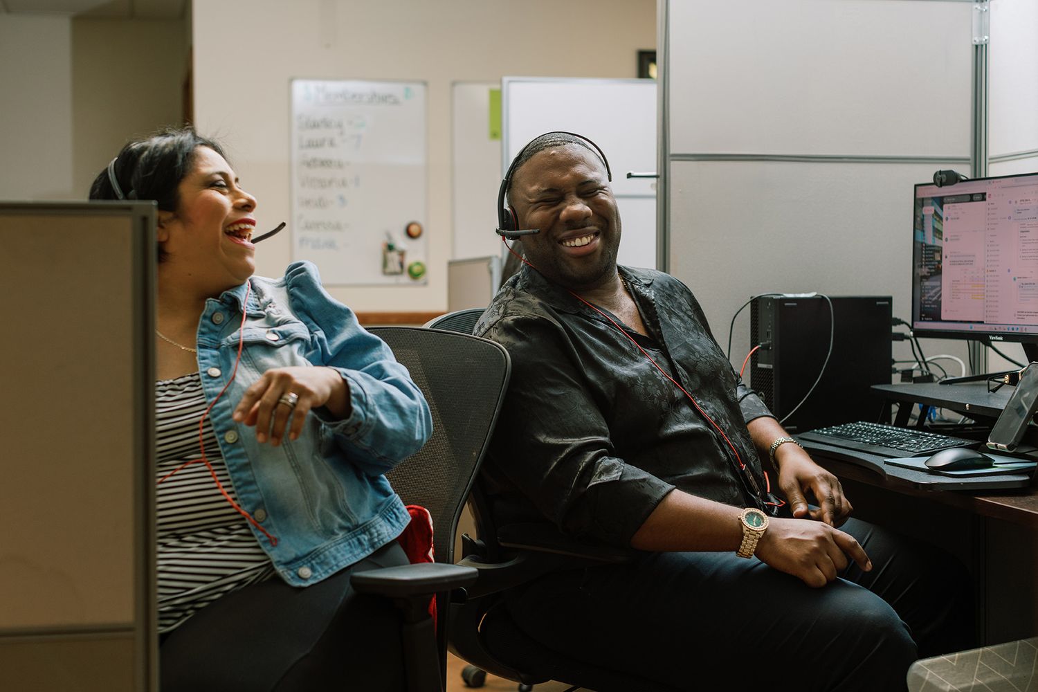 Two coworkers laugh together in an office cubicle, one wearing headphones.