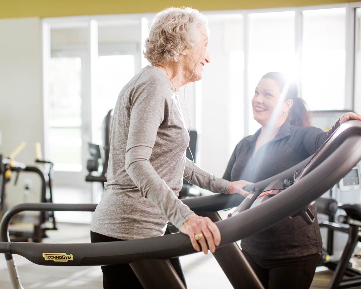 Elderly woman exercising on treadmill with smiling female trainer assisting her in a bright modern gym.