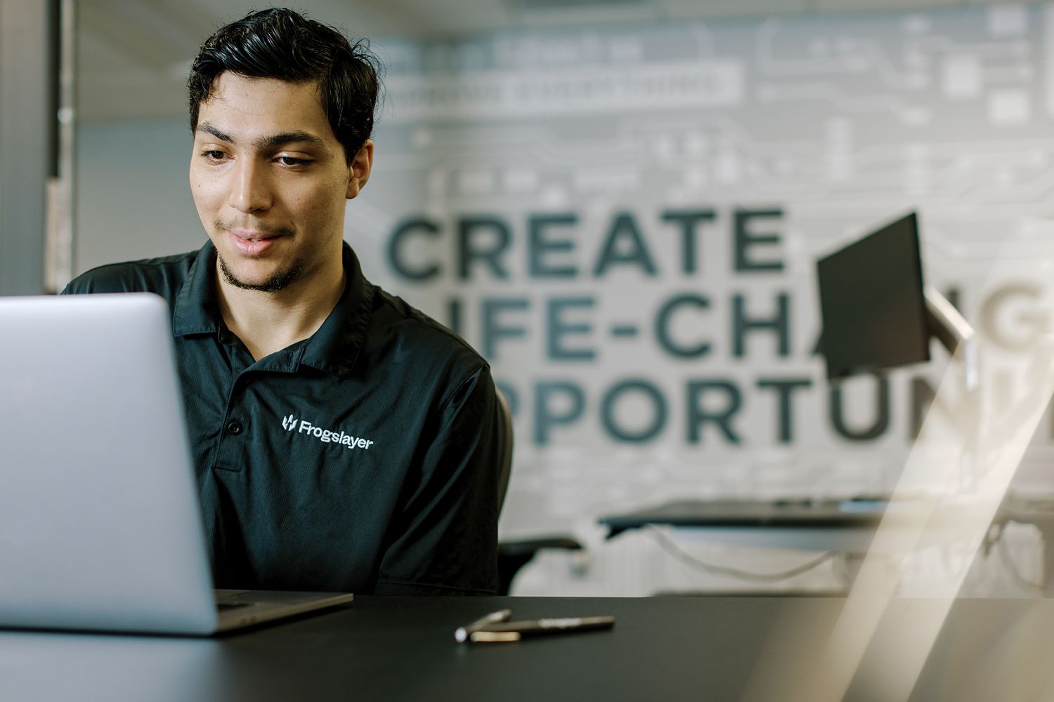 Young professional man working on laptop at desk with motivational quote on wall behind him.