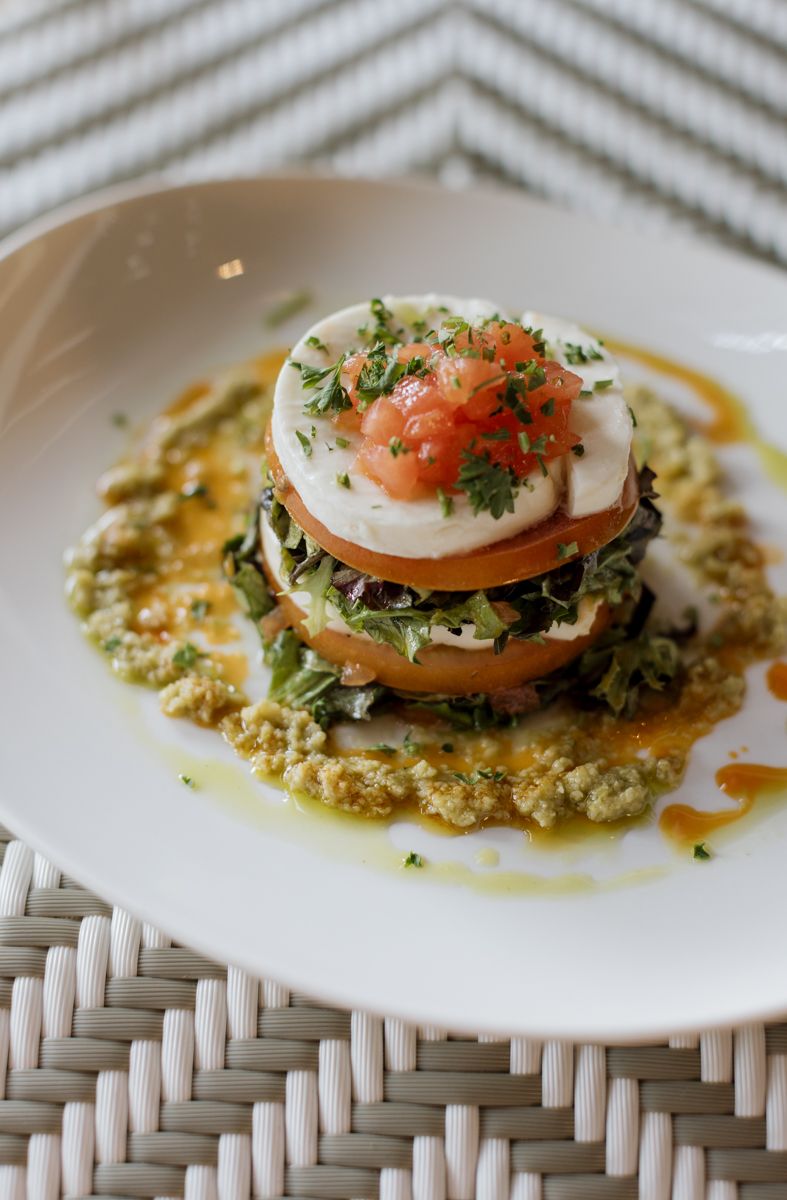 Elegant stacked caprese salad with poached egg, fresh greens, tomato, herbs, and pesto sauce on a white plate.