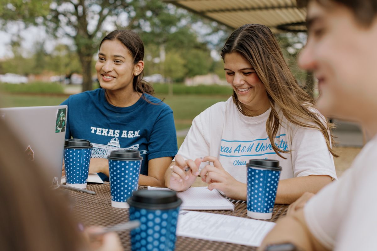 Two smiling female Texas A&M students sitting at an outdoor table with blue branded coffee cups on campus.