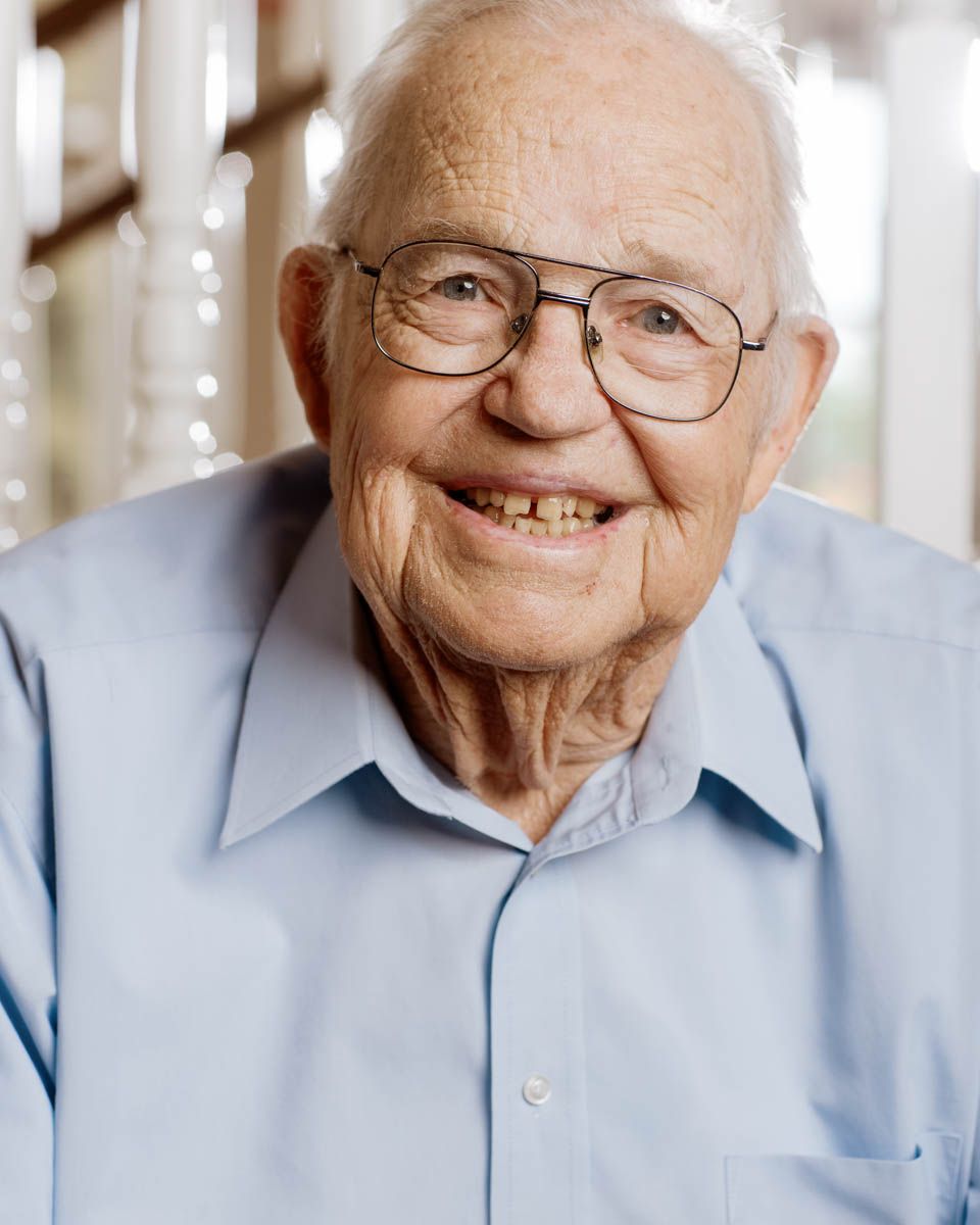 Cheerful elderly man with glasses wearing a light blue shirt smiling warmly outdoors.