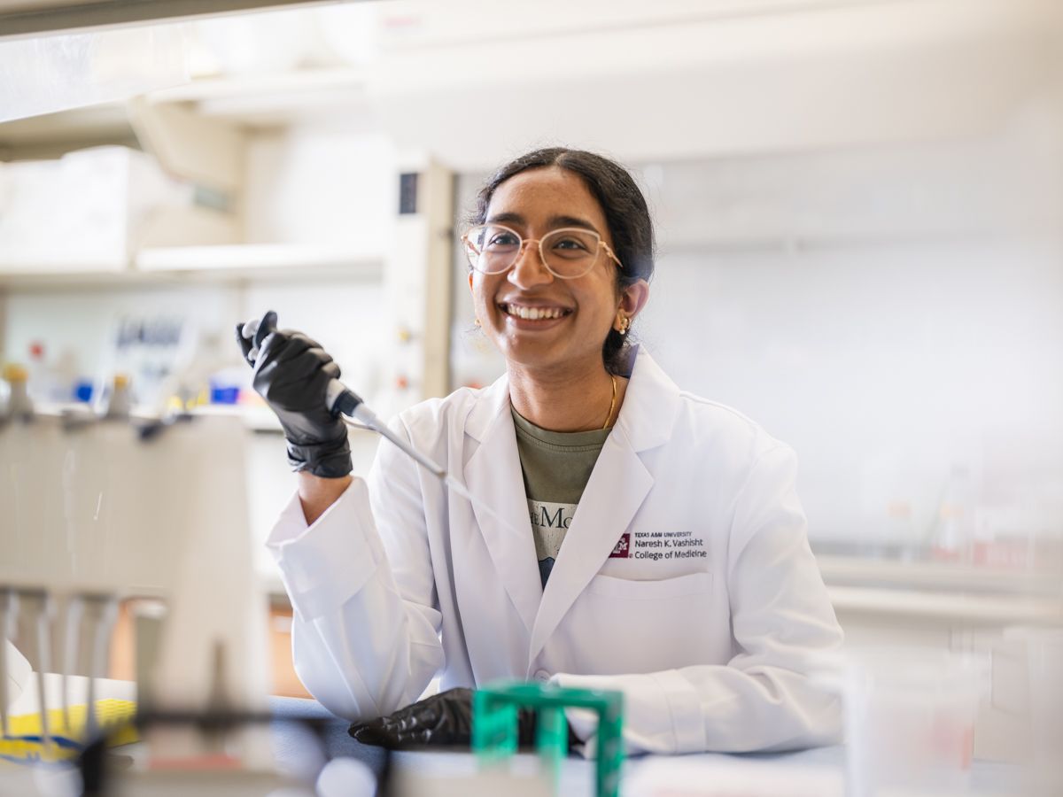 A smiling female scientist in a white lab coat uses a pipette in a bright research lab.