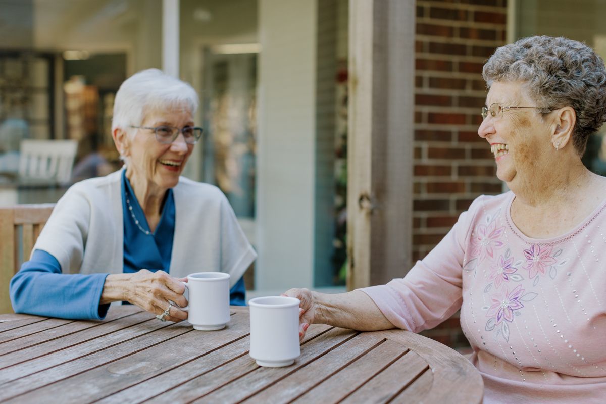 Two elderly women smile and chat over coffee cups at an outdoor wooden table at a senior living community.