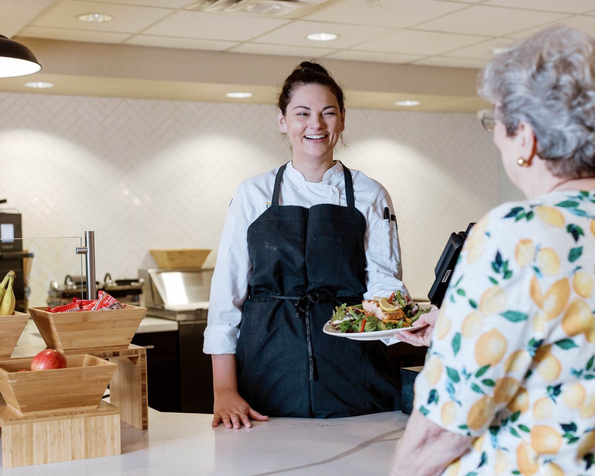 Smiling chef in black apron serving a fresh salad plate to a senior woman at a bright dining counter.