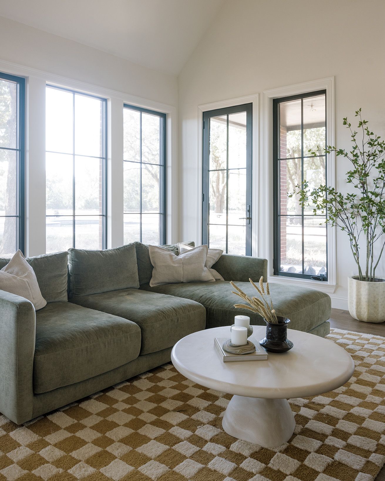 Sunlit living room with sage green sectional sofa, round white coffee table, and checkered wool rug.