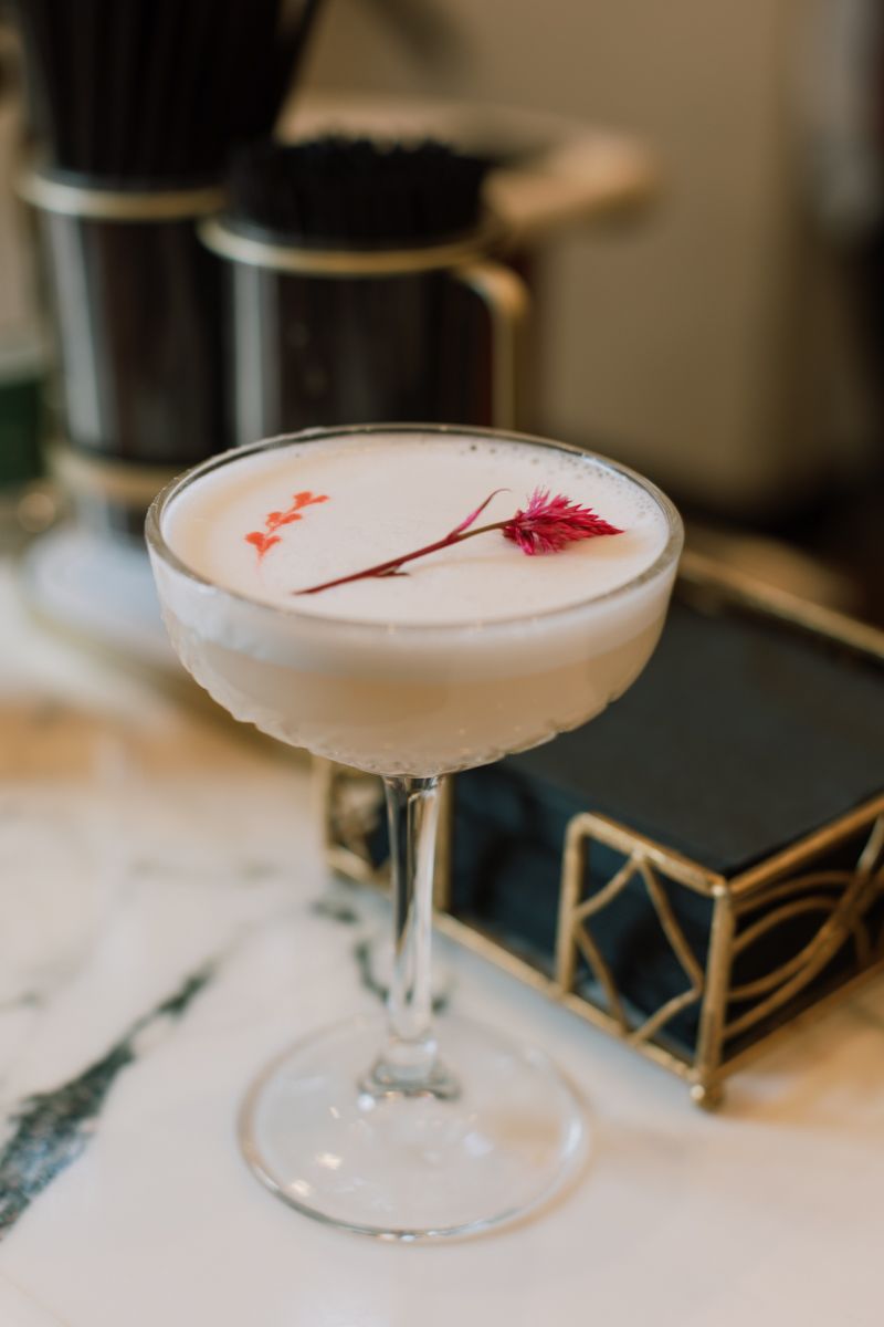 Elegant white foam cocktail garnished with a red botanical sprig served in a coupe glass on a marble bar.