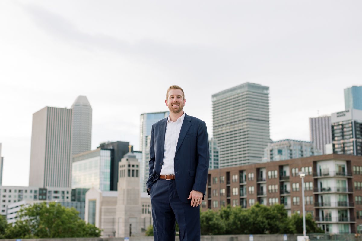 Professional man in navy suit stands outdoors with modern city skyline behind him.