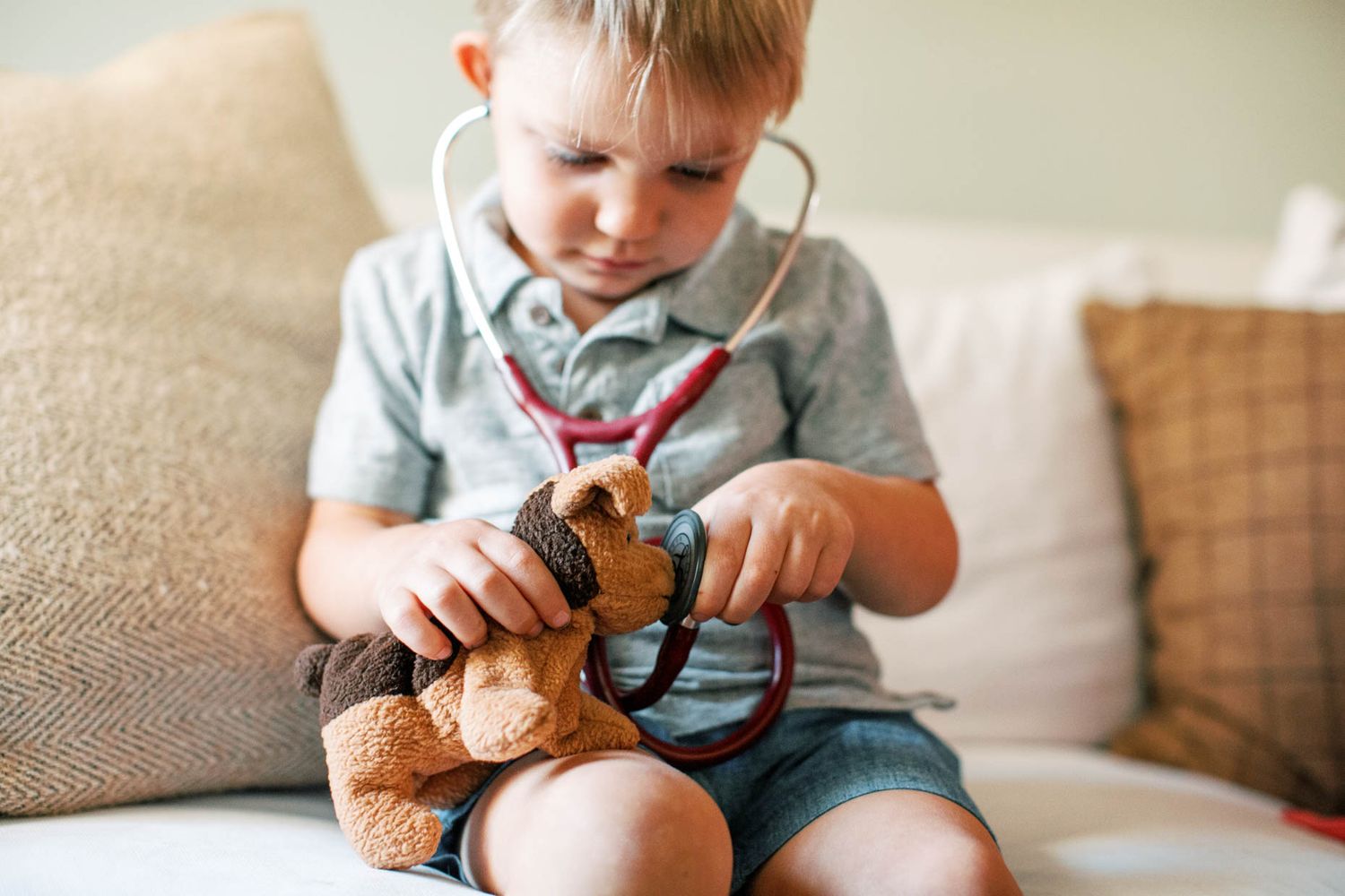 Young boy playing doctor with a teddy bear using a stethoscope while sitting on a couch.