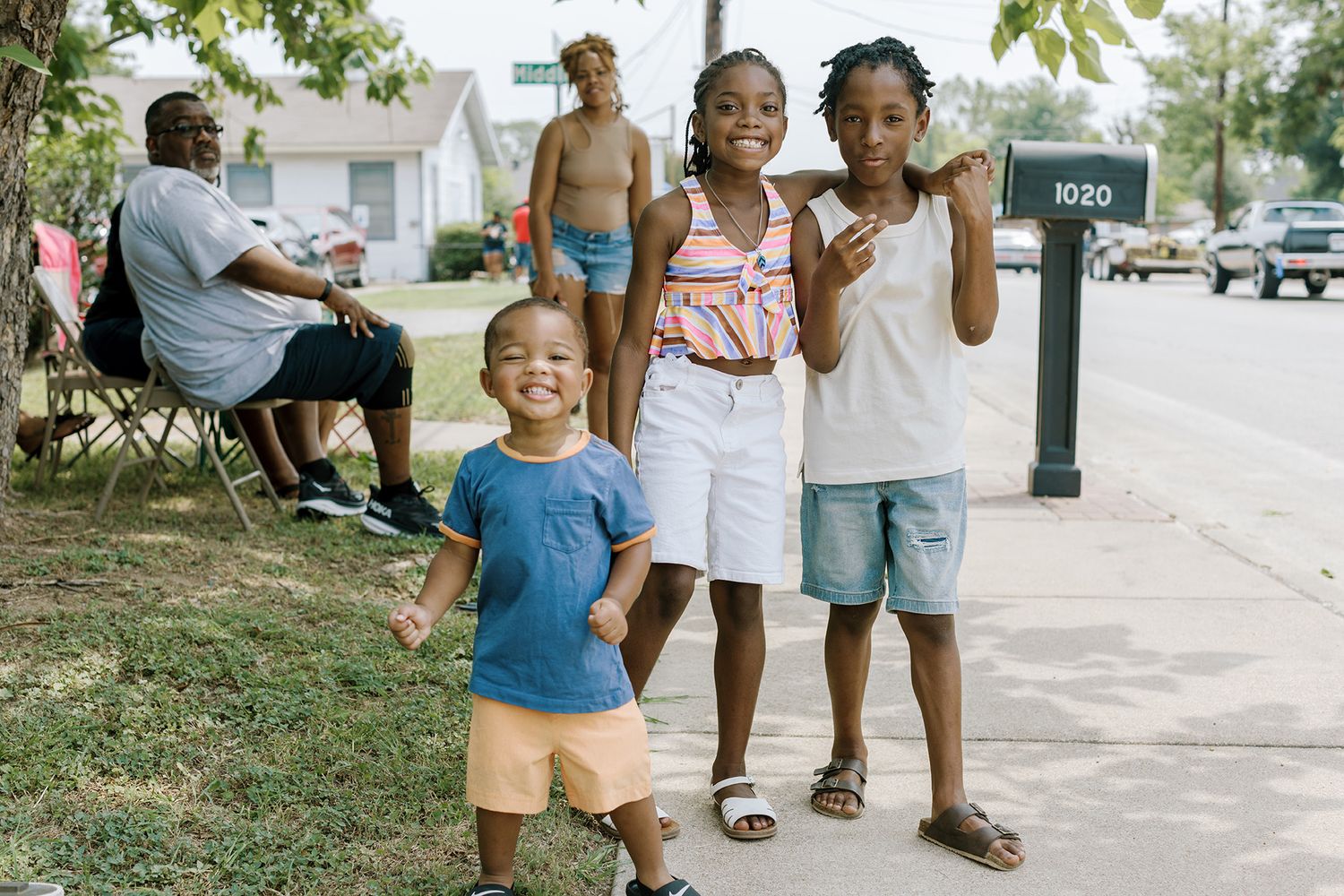 Three smiling Black children pose together on a sunny suburban sidewalk during a community event.