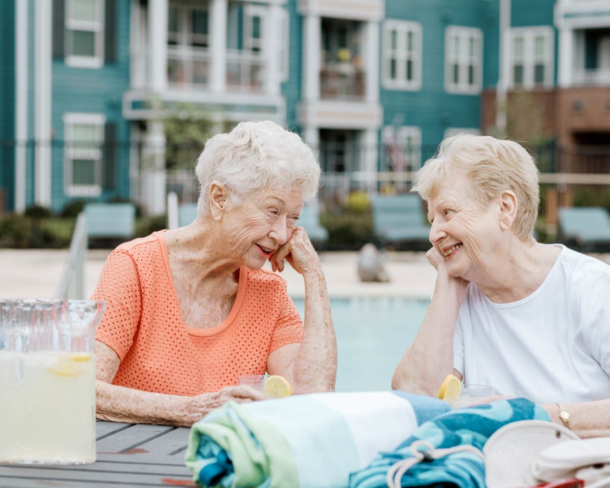 Two elderly women laughing together at an outdoor poolside table with lemonade on a sunny day.