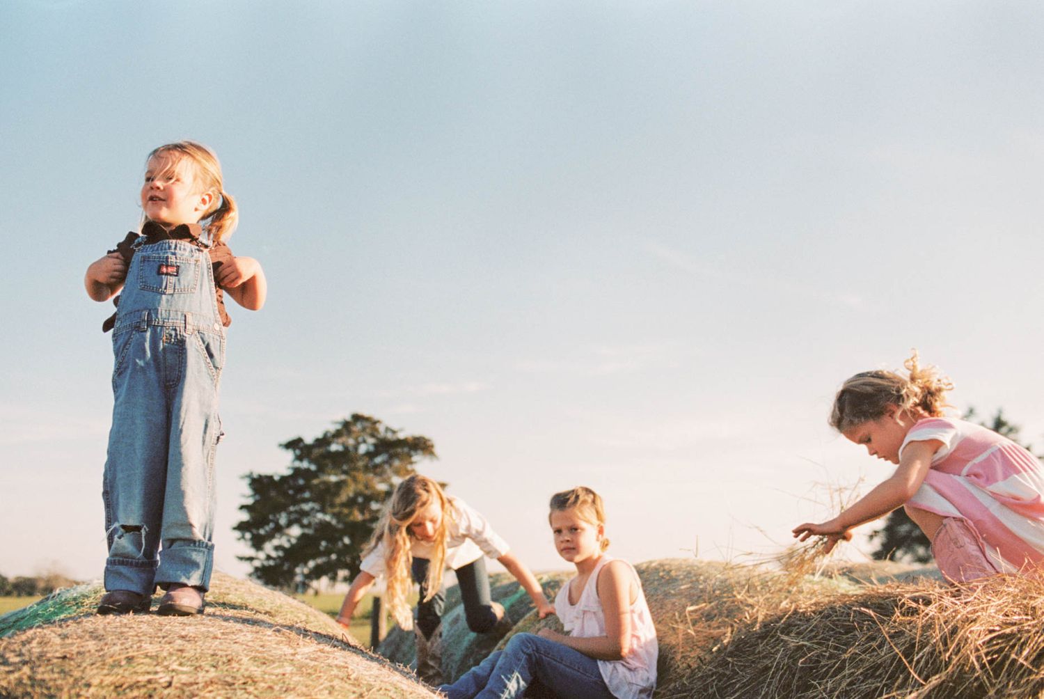 Children playing on a hay bale outdoors under a bright blue sky on a sunny day.