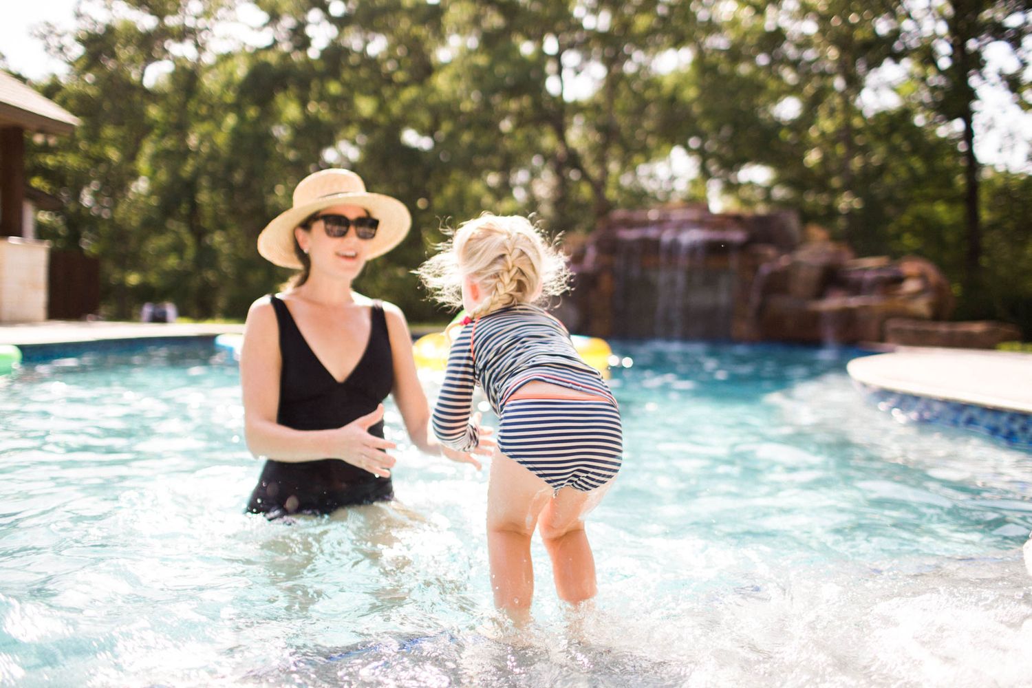Mother and toddler playing together in a backyard pool on a sunny day, with a waterfall feature in the background.