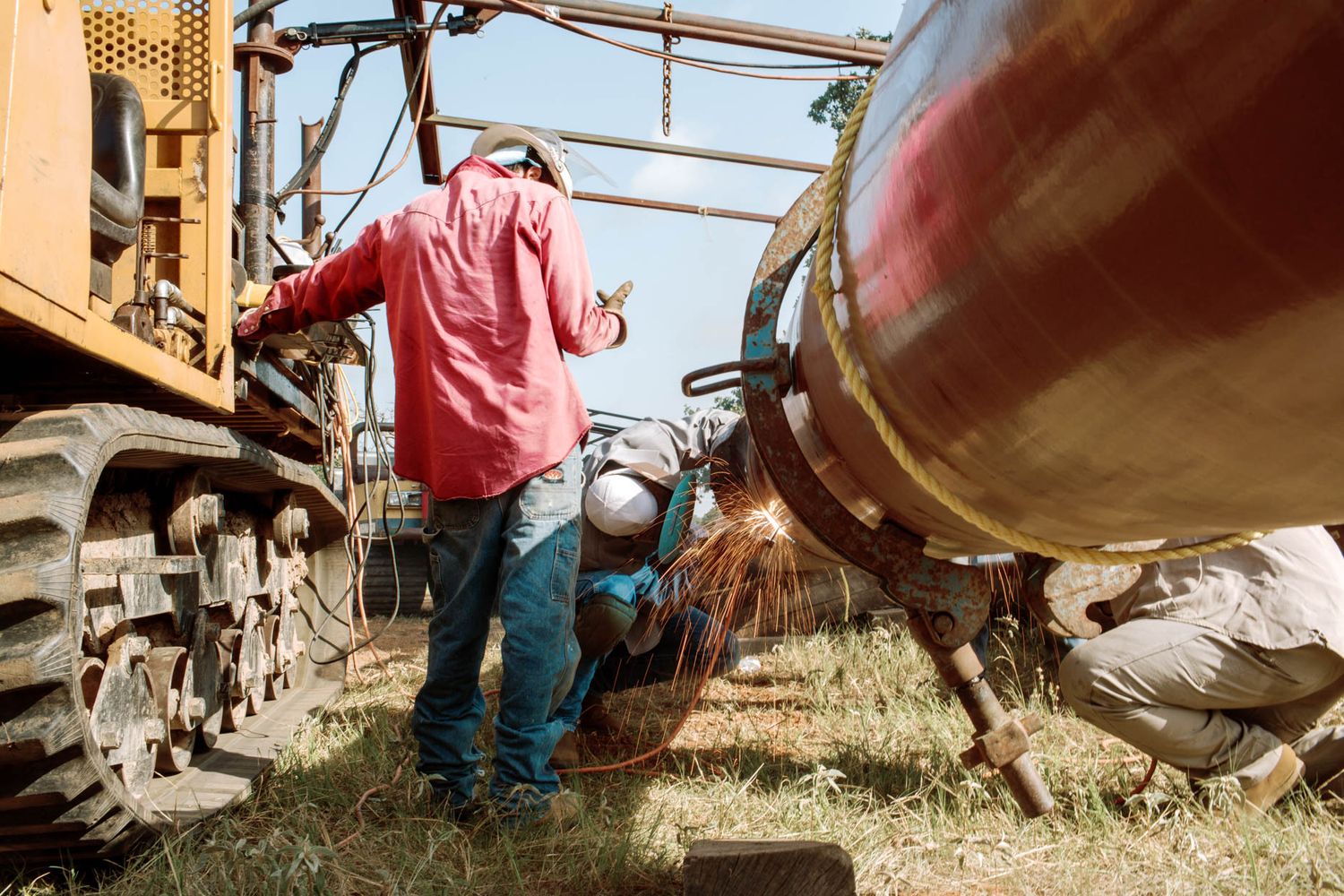 Workers welding a large pipeline near heavy construction equipment outdoors in a field.