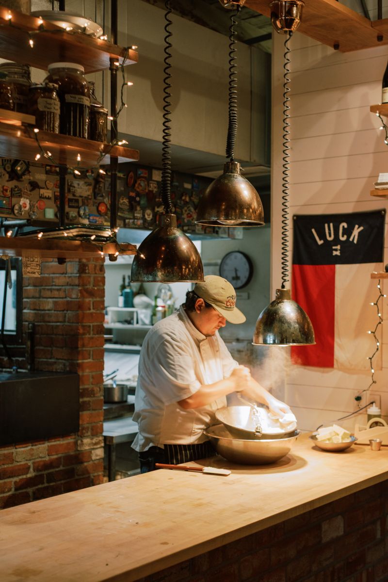 Chef preparing dough at a wooden counter in a warmly lit restaurant kitchen with hanging pendant lights.