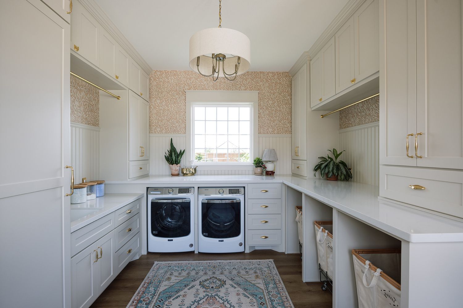 Bright laundry room with white cabinets, gold hardware, front-load washer and dryer, floral wallpaper, and a patterned area rug.