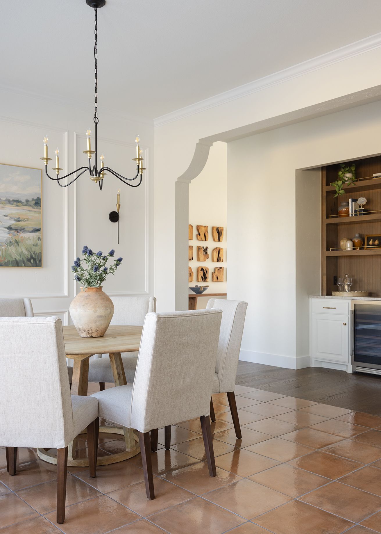 Bright dining room with round wood table, linen chairs, brass chandelier, terracotta tile floors, and built-in arched bookshelf.