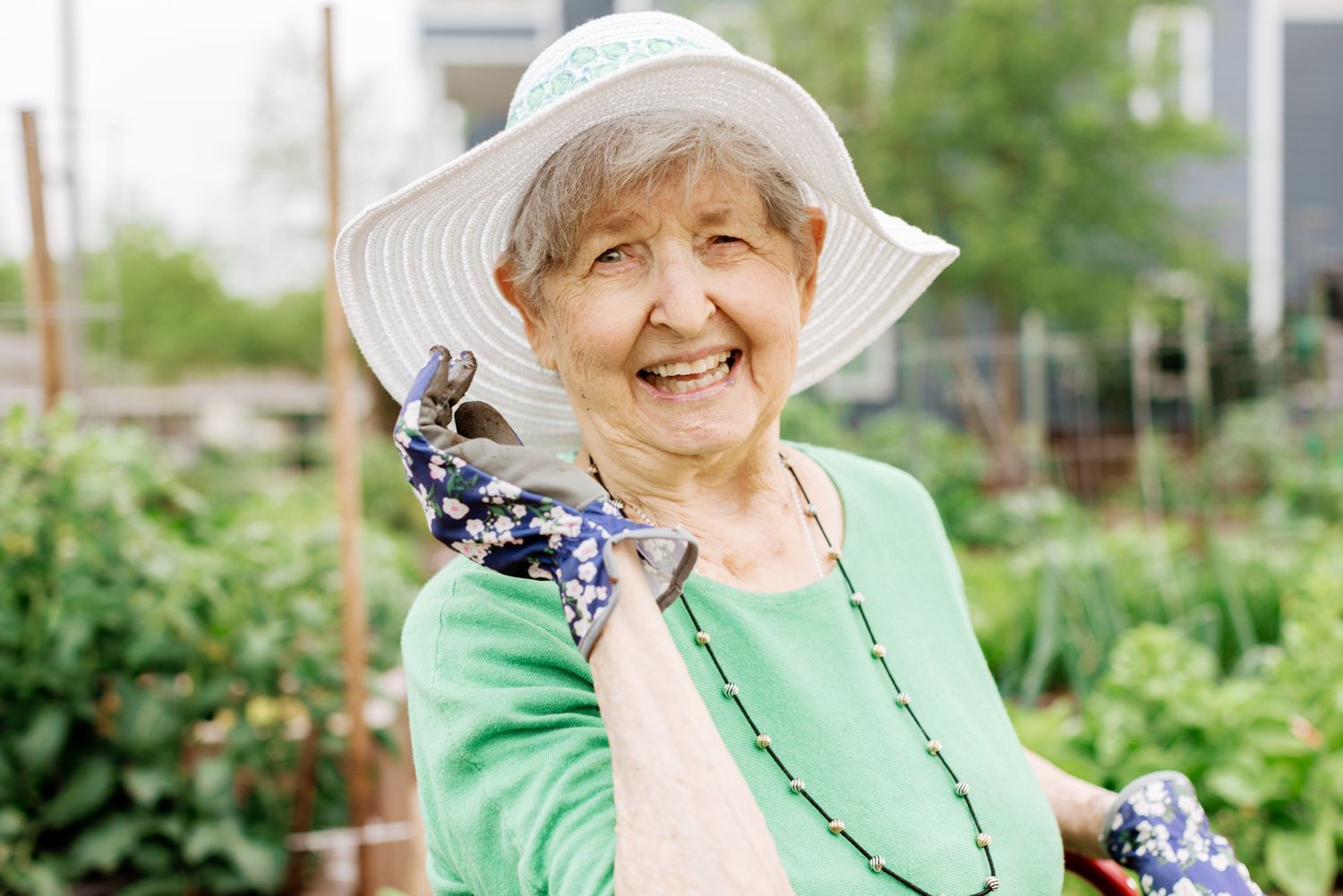 Happy senior woman in white hat and gardening gloves smiling in a lush community garden.