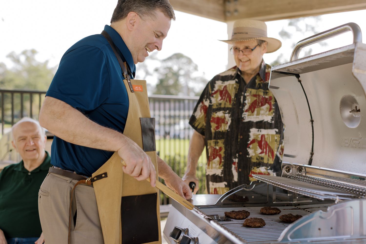 Two men smiling while grilling burgers outdoors on a gas grill at a senior living community cookout.