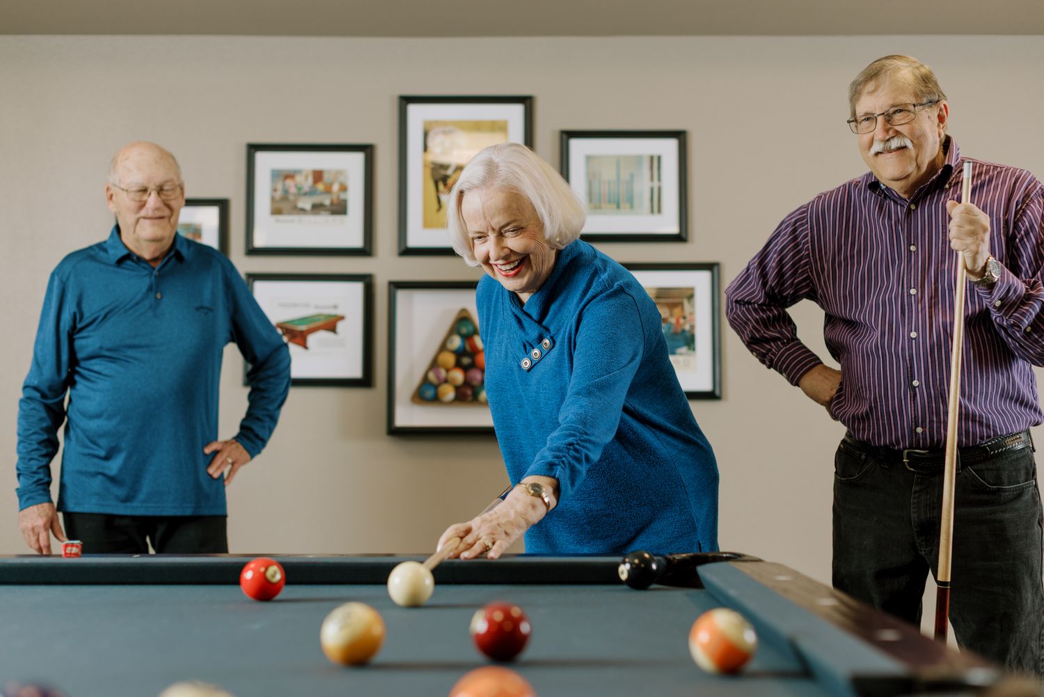 Three seniors enjoying a game of billiards in a bright recreation room with framed artwork on the wall.