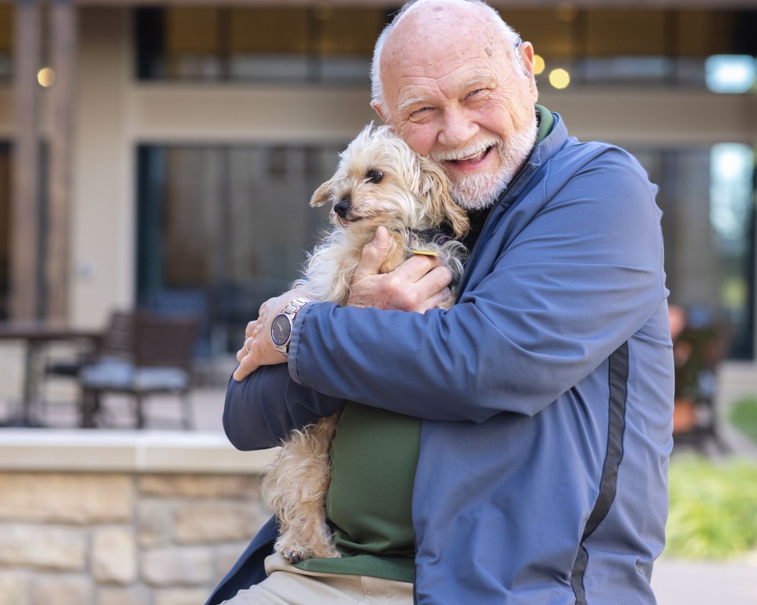 Smiling senior man with white beard hugging a small fluffy terrier dog outdoors near a building.
