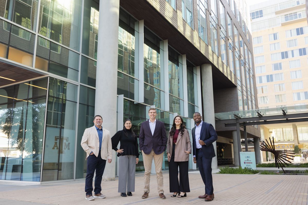 Five diverse business professionals pose together outside a modern glass building.