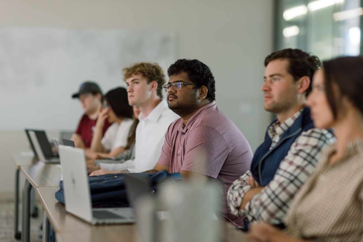 Diverse group of students listening attentively in a modern classroom with laptops.