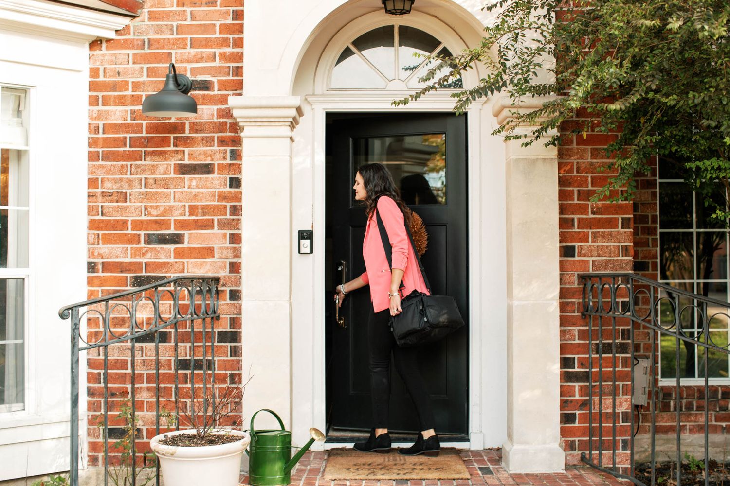 Woman in pink blazer and black dress entering a brick home through a black front door.