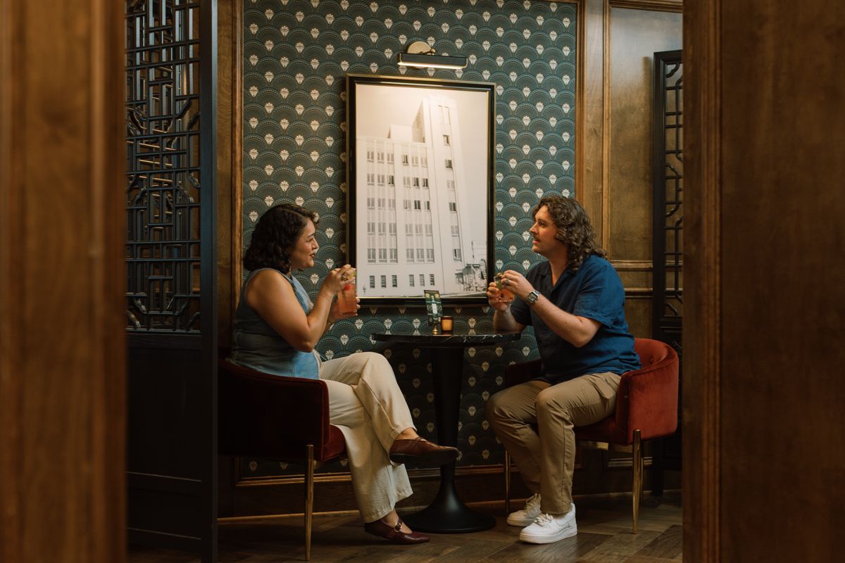 Two people toasting drinks at a cozy restaurant table with teal wallpaper and elegant decor.
