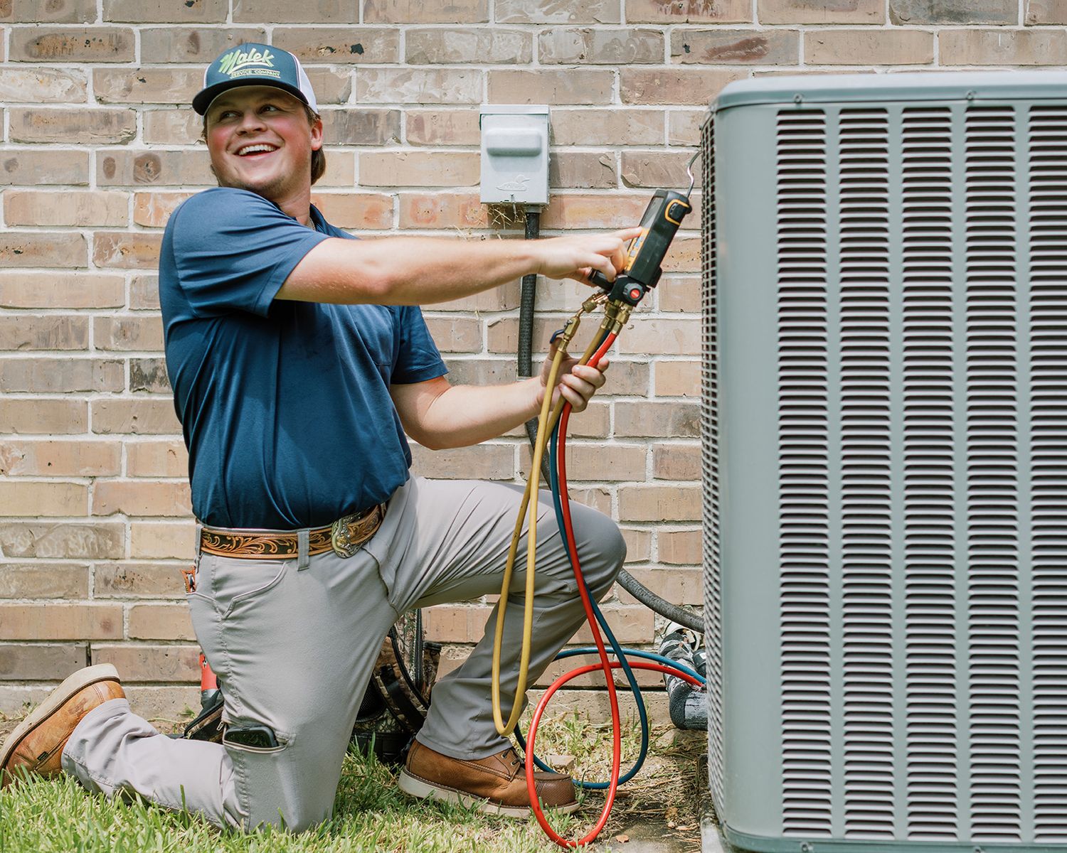HVAC technician kneeling outdoors, smiling while servicing an air conditioning unit with refrigerant gauges.