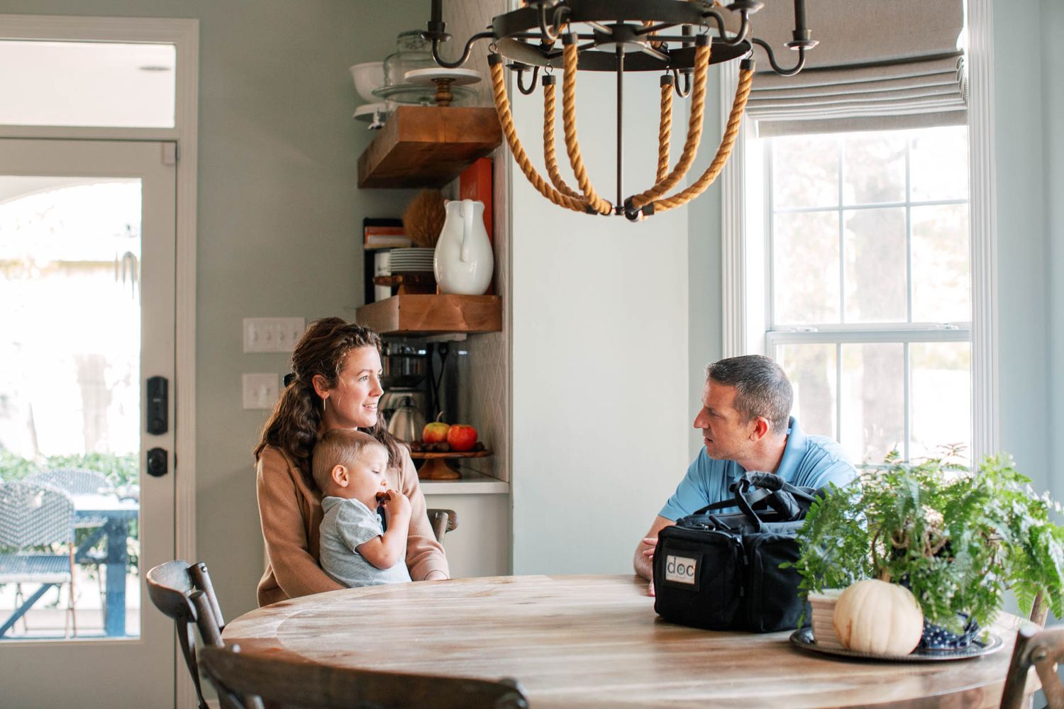 Mother holding infant meets with doctor at round dining table in bright home kitchen setting.