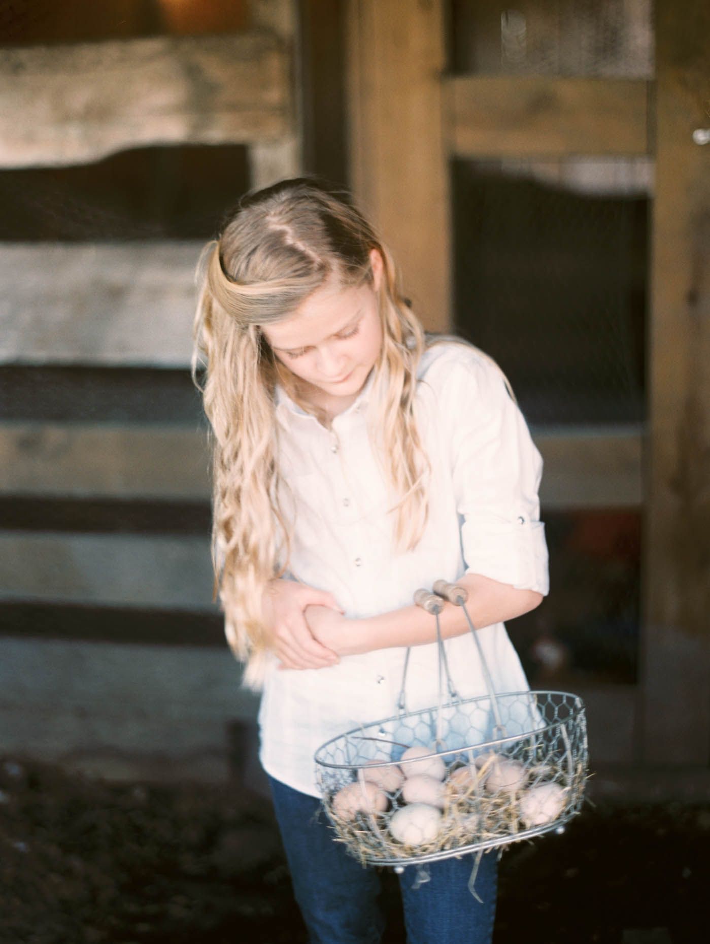 Young girl holding a wire basket filled with fresh farm eggs outside a wooden barn.