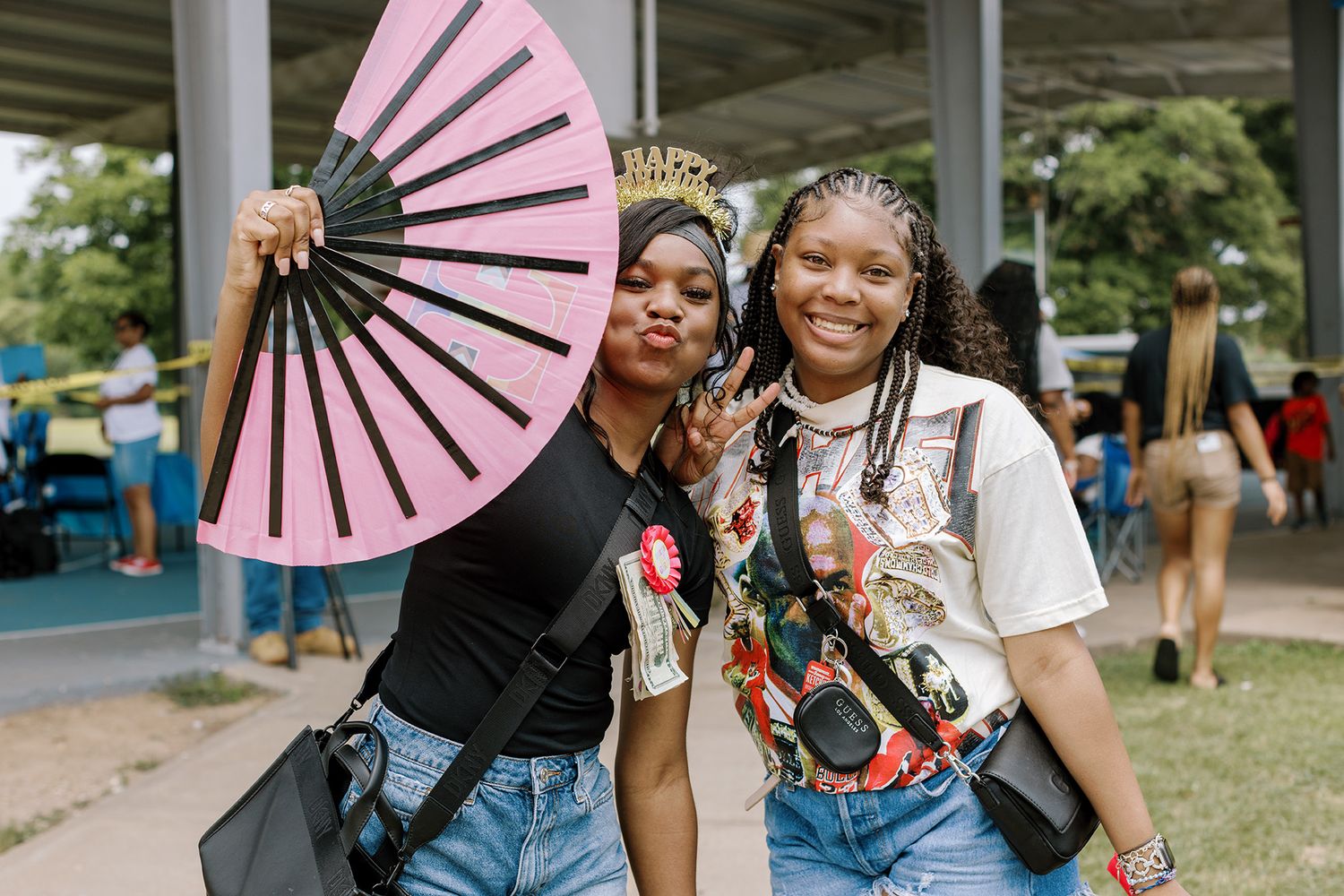 Two young women posing outdoors at a festival, one holding a large pink decorative fan.