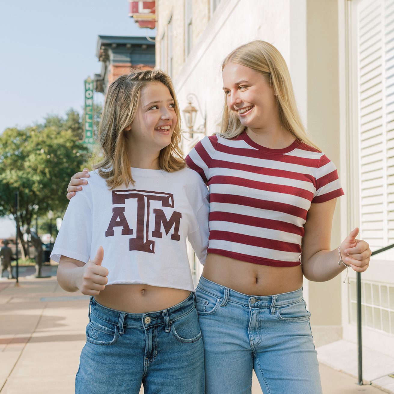Two smiling young women in Texas A&M and striped crop tops give thumbs up on a sunny sidewalk.