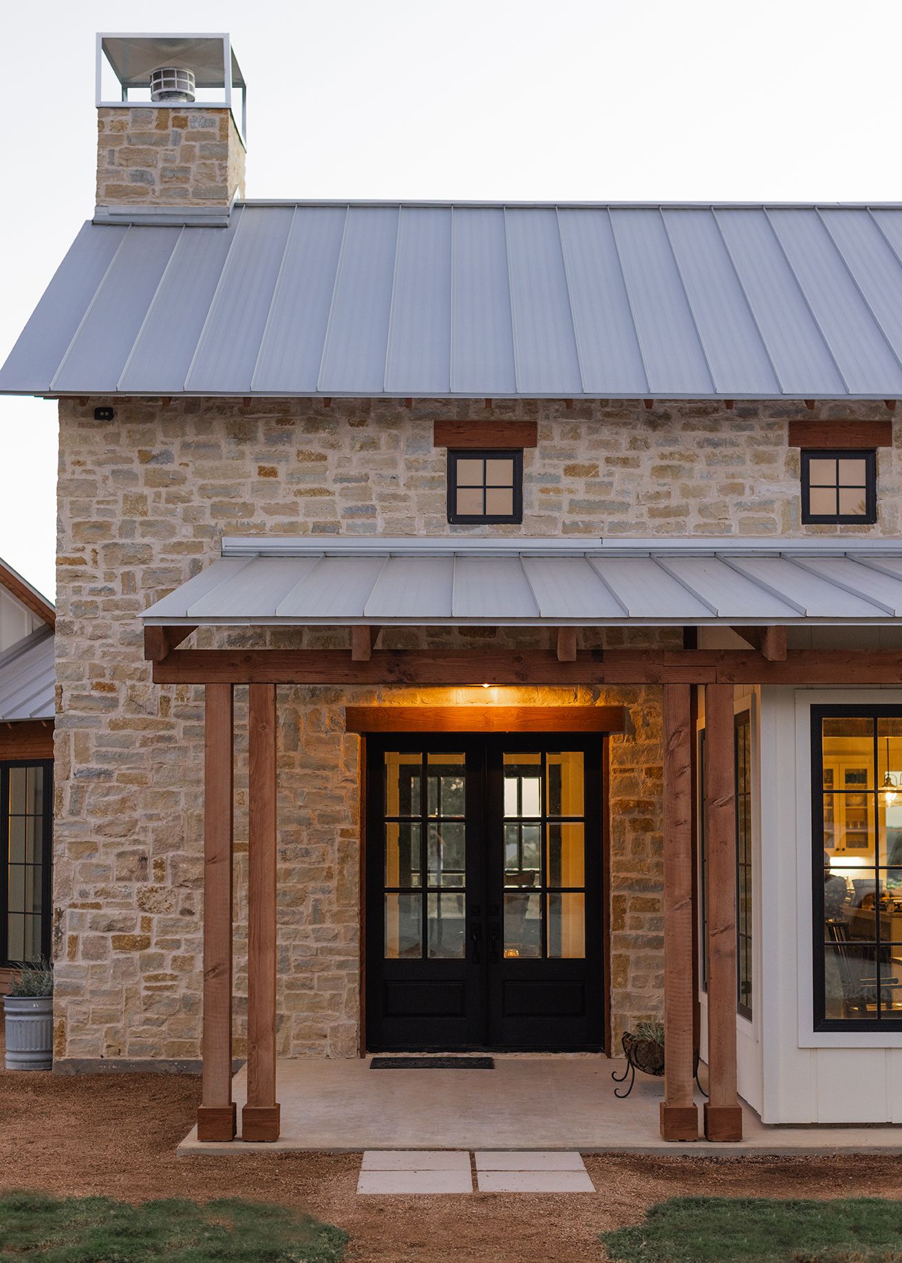 Rustic stone farmhouse exterior at dusk with metal roof, wood porch beams, black French doors, and warm interior lighting.