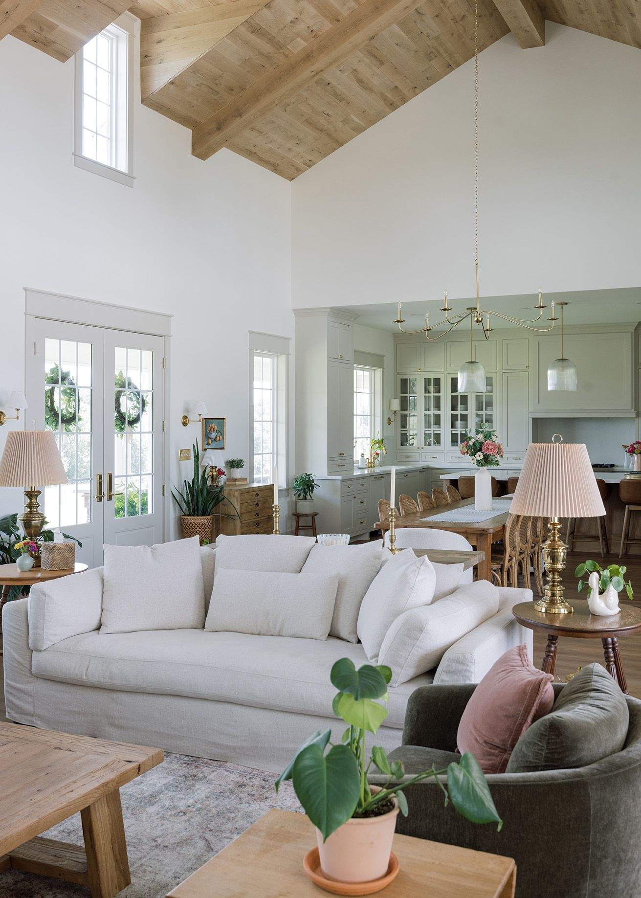Bright open-concept living room with vaulted wood beam ceiling, white sofa, and view into sage green kitchen.
