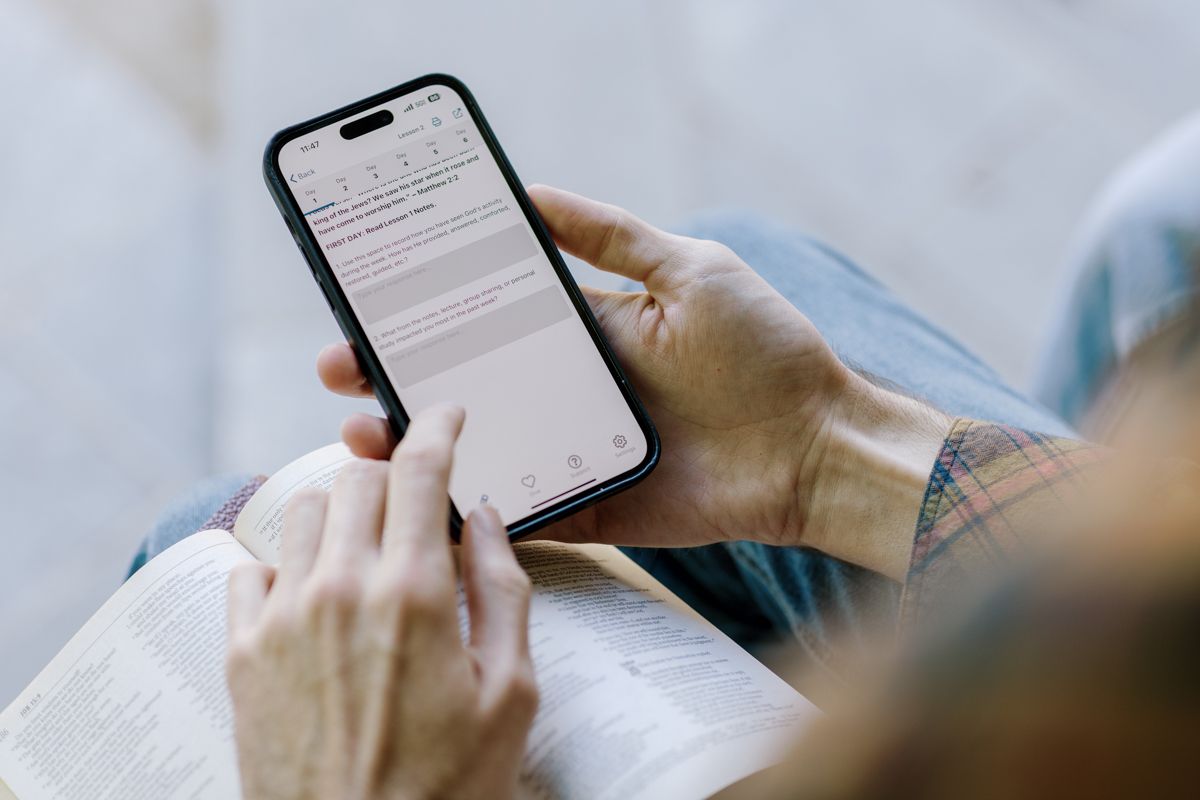 Person holding smartphone while reading, with an open book on their lap outdoors.