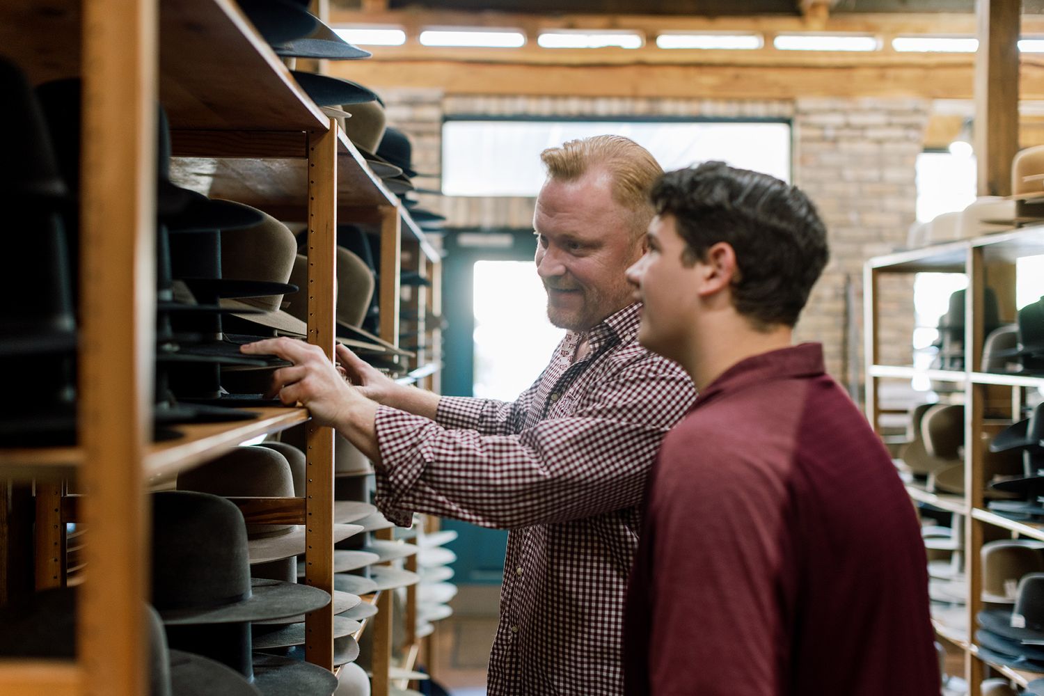 Two men smiling while browsing hats on wooden shelves in a hat shop.