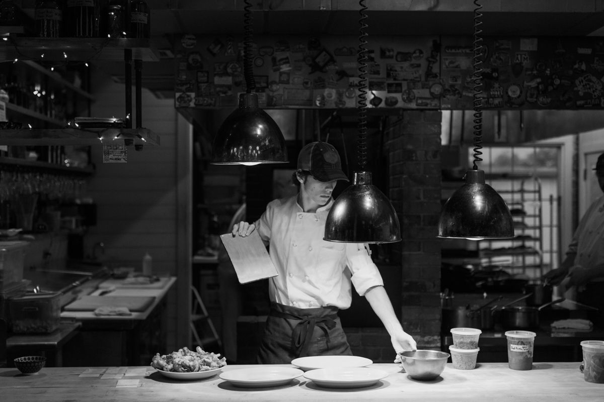 Black and white photo of a chef preparing plates in a dimly lit restaurant kitchen.