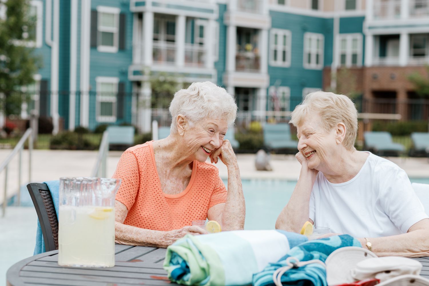Two senior women laughing together at outdoor table near a resort-style swimming pool.