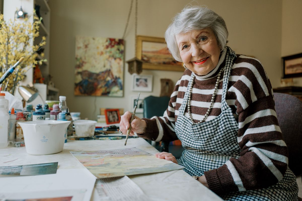 Elegant senior woman smiling while painting watercolors at a desk in her art-filled studio.